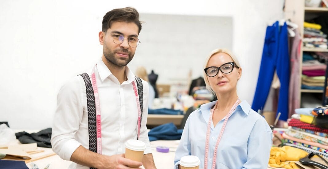 Two fashion designers or tailors standing in a clothing workshop, holding coffee cups, with measuring tapes around their necks. The background shows shelves filled with fabrics and sewing supplies.