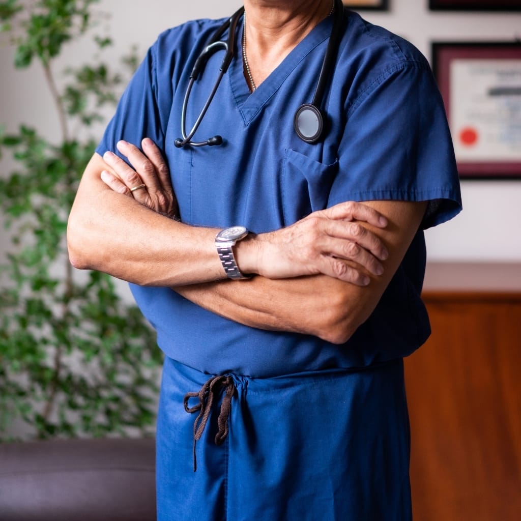 A close-up of a healthcare professional wearing blue scrubs with arms crossed, a stethoscope around their neck, and a watch on their wrist. Part of a framed certificate or diploma is visible in the background.