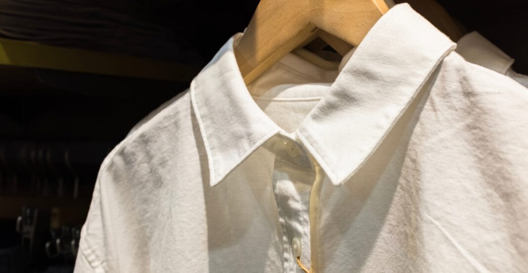 A close-up of a white collared shirt hanging on a wooden hanger, with the top button fastened and the collar neatly prominent.