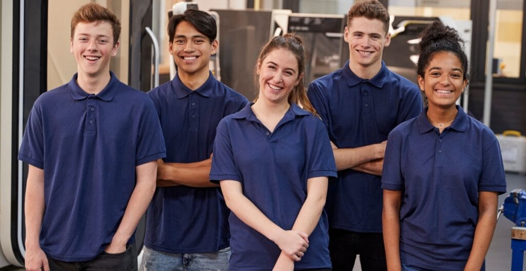 "Five teenagers standing together in blue polo shirts, smiling in what appears to be a workshop or school lab."