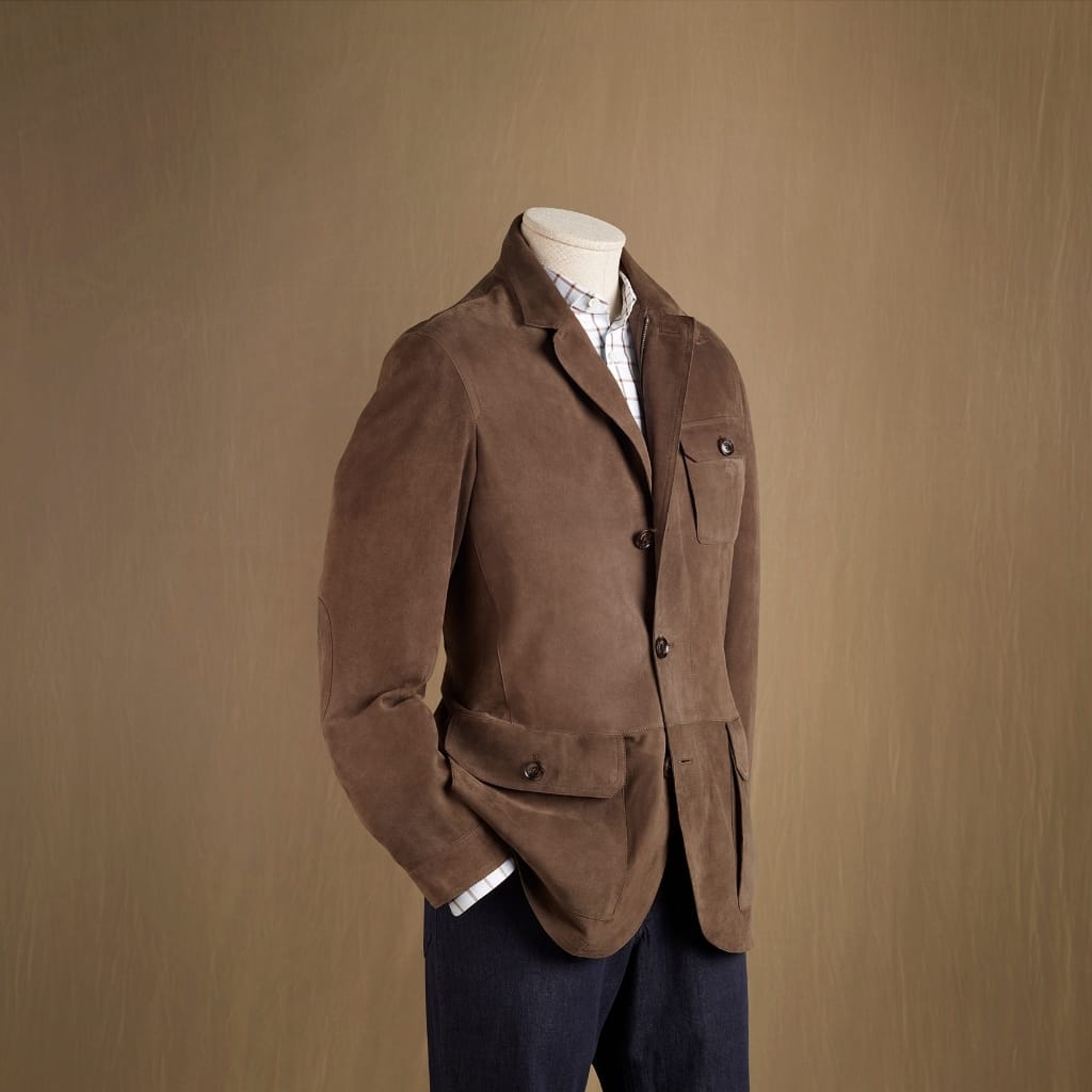 A headless mannequin dressed in a brown suede blazer over a light checkered shirt, with dark trousers, posed against a warm brown backdrop.