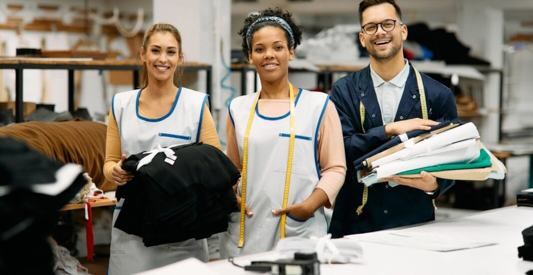 Three smiling people, two women and one man, standing in a workspace or sewing shop. The women are wearing work aprons with measuring tapes, one holding a stack of black fabric, and the man is holding a pile of fabric and garment pieces. Shelves with fabric and sewing materials are visible in the background.