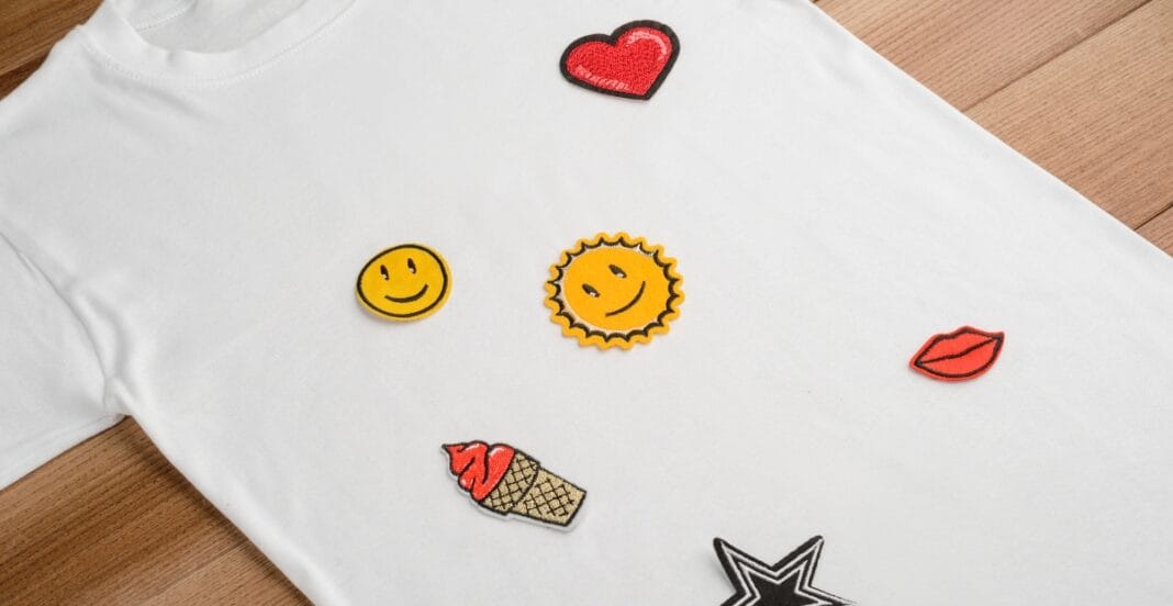 A white T-shirt laid flat on a wooden surface with various colorful embroidered patches scattered on it, including a red heart, a yellow smiley face, a sun, red lips, an ice cream cone, and a black-and-white star.