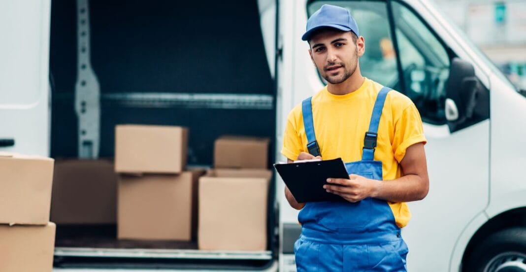 A young male delivery worker wearing a yellow shirt and blue overalls stands beside an open delivery van with several cardboard boxes inside. He holds a clipboard and looks at the camera.