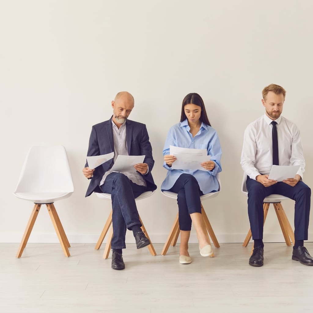Three people sitting on chairs against a plain white wall. The first is an older man with a beard in a dark blazer and jeans, looking at papers. The second is a young woman with long dark hair, wearing a light blue shirt and dark pants, also reading papers. The third is a young man with short blond hair, dressed in a white shirt and dark tie, holding papers and looking down.