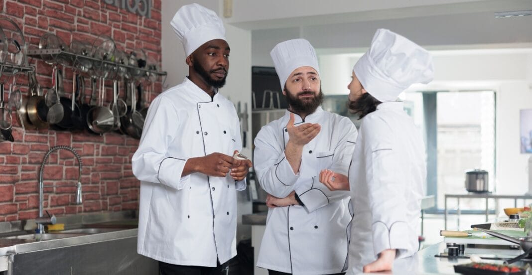 Three chefs in white uniforms and tall hats are engaged in a discussion in a commercial kitchen. Two men and one woman listen intently, with one man gesturing as he speaks. The kitchen features hanging pots and pans on a brick wall.