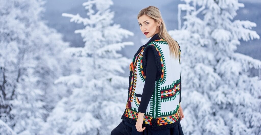 A young woman with blonde hair wearing a colorful, embroidered vest over a black outfit, standing outdoors in a snowy winter landscape with snow-covered trees in the background.