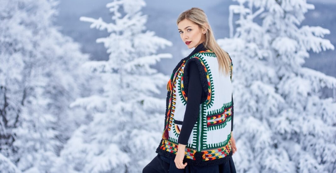 A young woman with blonde hair wearing a colorful, embroidered vest over a black outfit, standing outdoors in a snowy winter landscape with snow-covered trees in the background.
