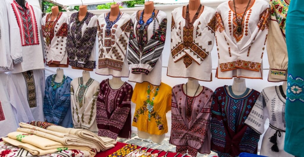 A display of traditional embroidered clothing on mannequins at an outdoor market stall. The garments feature intricate patterns in various colors, primarily white, with some in yellow and maroon, alongside folded embroidered textiles on the table.