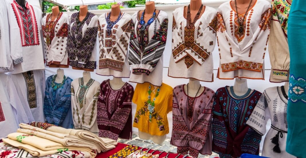 A display of traditional embroidered clothing on mannequins at an outdoor market stall. The garments feature intricate patterns in various colors, primarily white, with some in yellow and maroon, alongside folded embroidered textiles on the table.