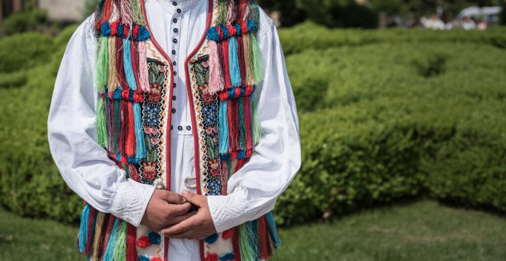 A person standing outdoors wearing a traditional, colorful embroidered garment with multicolored fringes and details on the vest, over a white shirt.