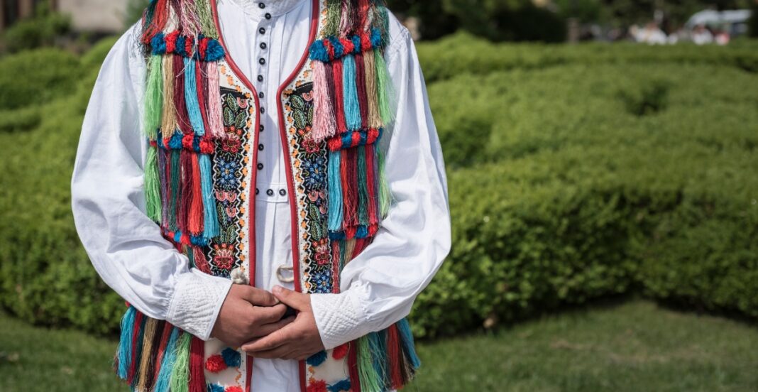 A person standing outdoors wearing a traditional, colorful embroidered garment with multicolored fringes and details on the vest, over a white shirt.