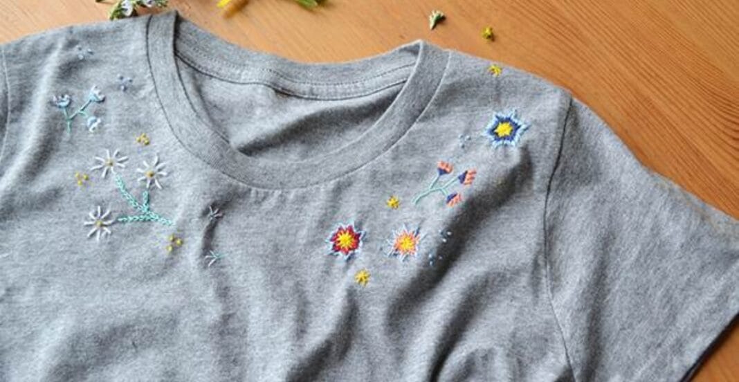 A gray T-shirt with colorful embroidered floral and star designs near the shoulder and upper chest area, placed on a wooden surface.