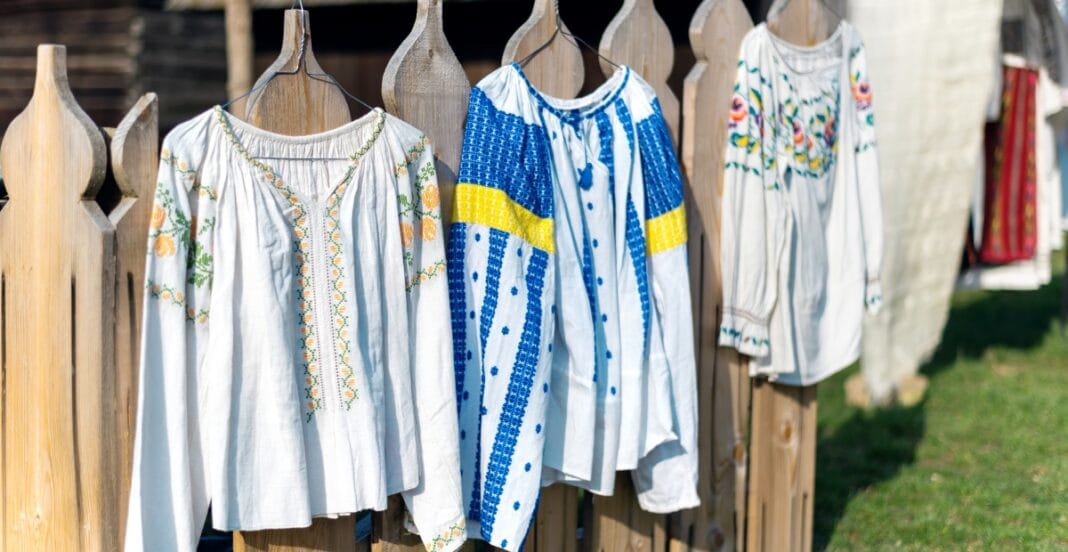 Three embroidered and patterned traditional blouses hanging on a wooden fence outdoors, with grass visible in the background.