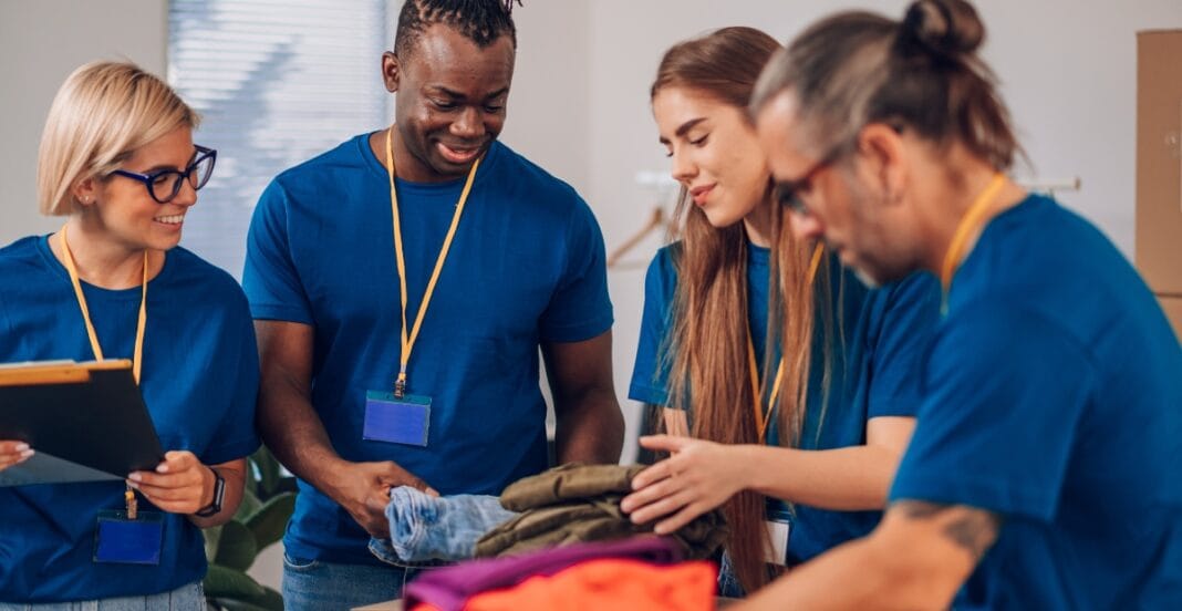 A diverse group of four young adults wearing blue shirts and name badges stands around a table, sorting through a pile of clothes and smiling as they work together.