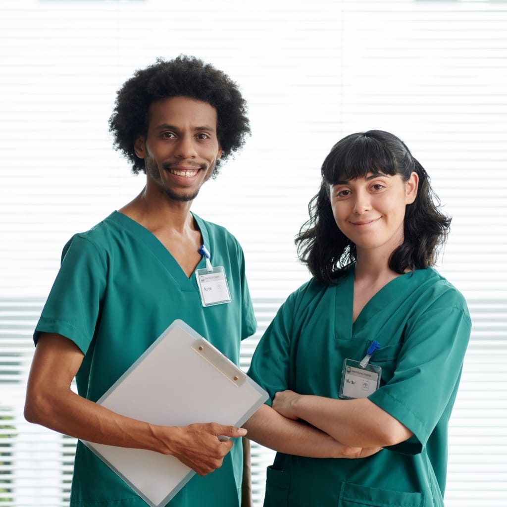 Two healthcare professionals, a man and a woman, dressed in green scrubs, standing side by side with the man holding a clipboard and both smiling confidently in front of a window with blinds.