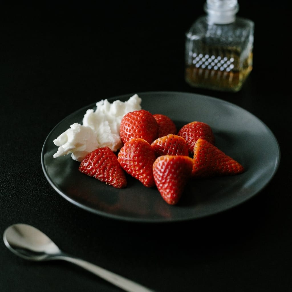 A black plate with fresh strawberries and a dollop of whipped cream, set on a dark surface with a spoon nearby and a glass bottle in the background.