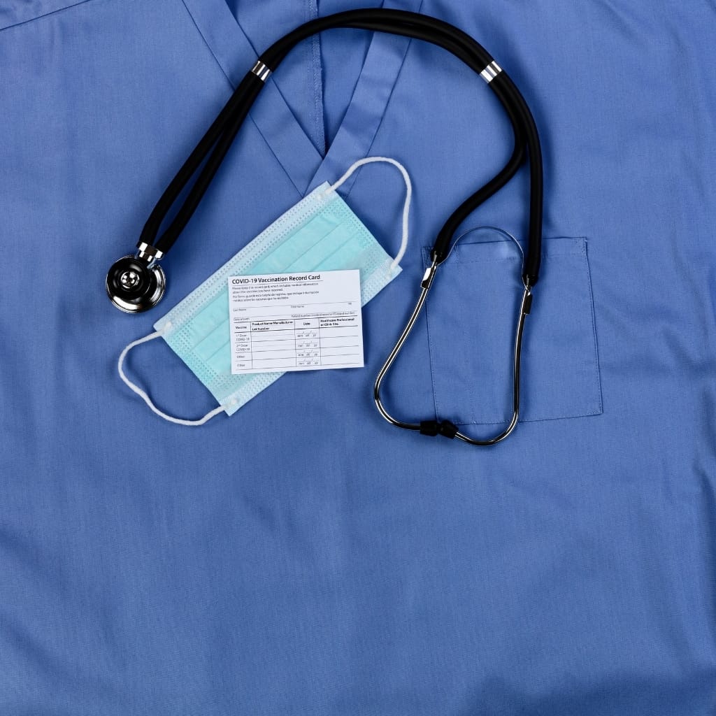 A close-up of a blue medical scrubs fabric featuring a stethoscope, a face mask, and a COVID-19 vaccination record card laid out on top.