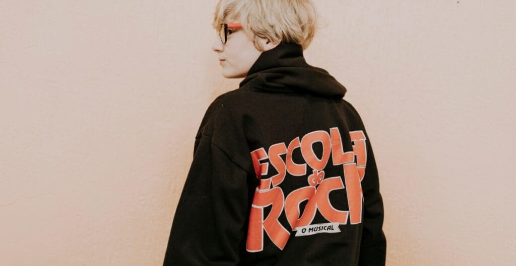 A person with blond hair wearing a black hoodie with large red and white text on the back, standing sideways against a pale wall.