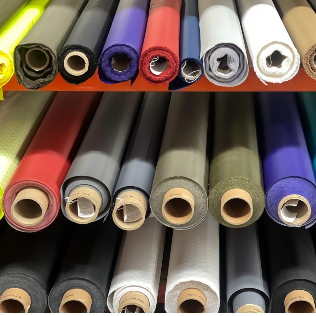 Shelves filled with rolled-up rolls of vinyl or fabric material in various colors, including bright yellow, black, purple, red, white, gray, beige, and green.