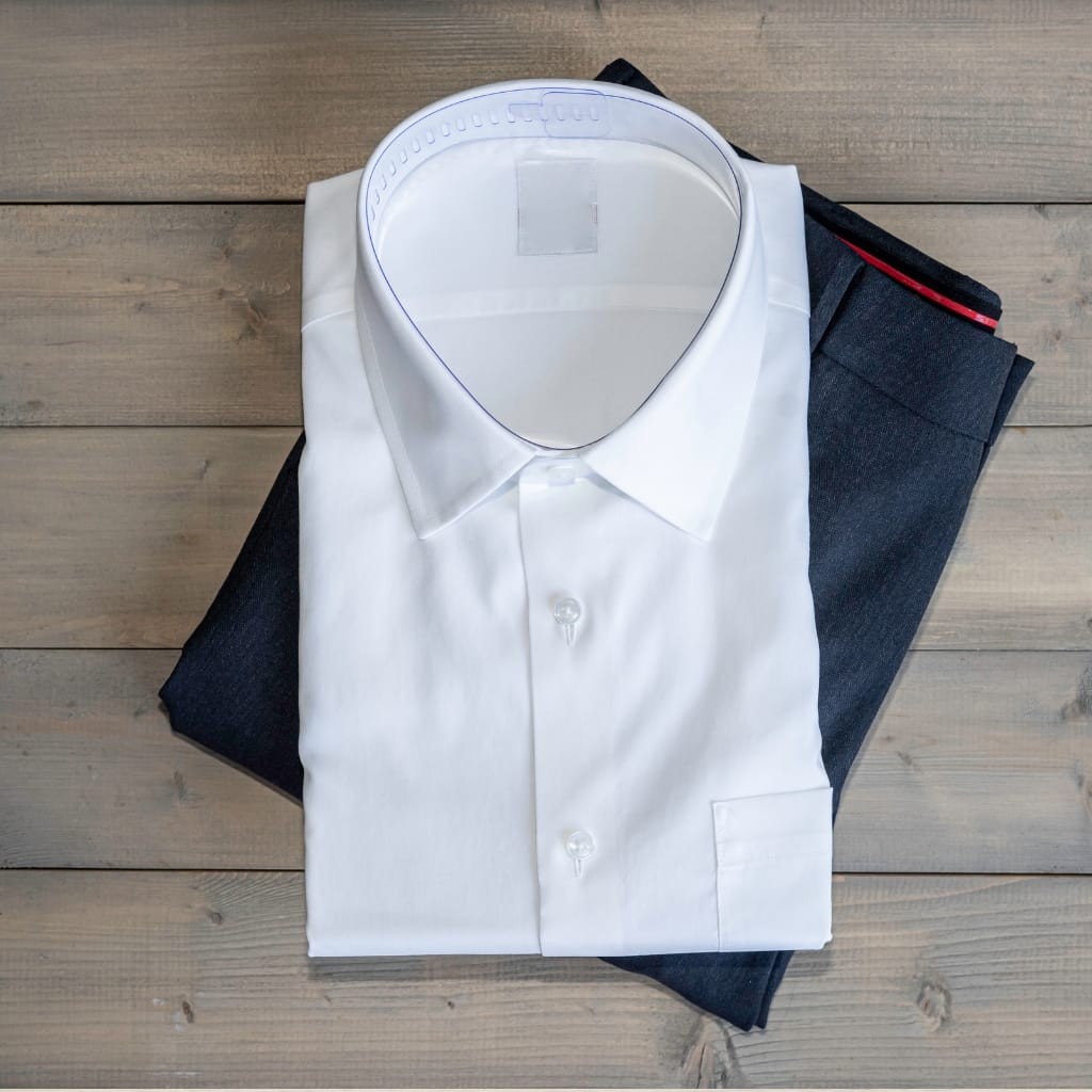 A neatly pressed white dress shirt folded on top of a pair of dark trousers, viewed from above. The shirt is buttoned and the collar is upright.