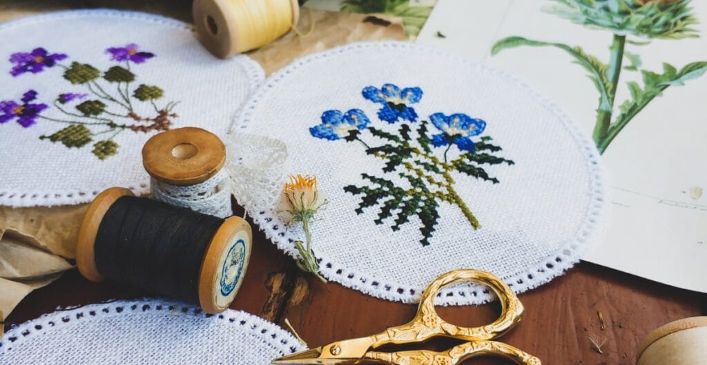 A close-up view of embroidery materials on a wooden surface, including finished and in-progress embroidered rounds with floral designs, spools of black and white thread, a pair of golden scissors, a flower, and a piece of paper with a botanical illustration.