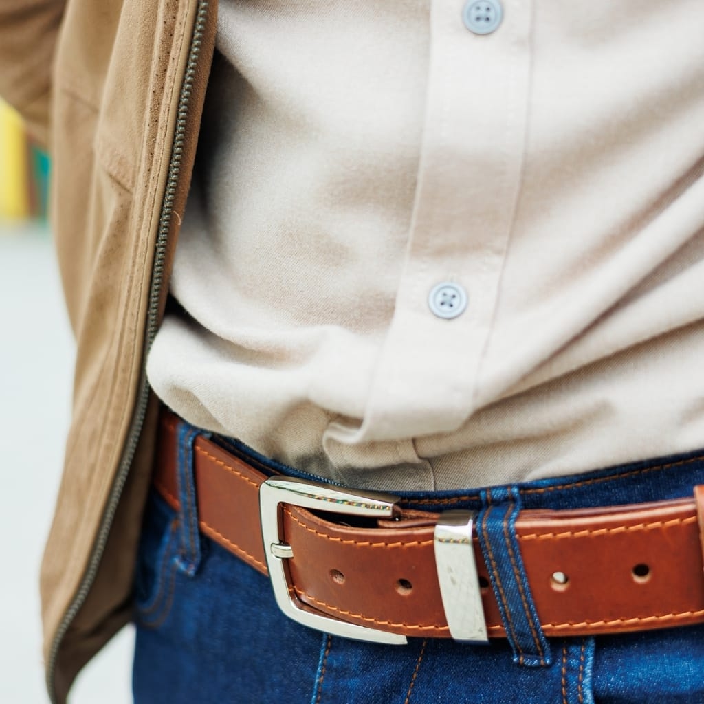 A close-up of a person's midsection showing a tan suede jacket, beige button-down shirt, a brown leather belt, and blue jeans.