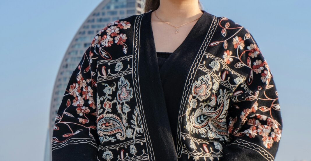 A person wearing a black embroidered jacket with colorful floral and abstract patterns, standing outdoors with a modern building in the background.