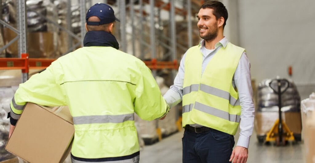 Two men in a warehouse shaking hands. Both are wearing high-visibility jackets; one is holding a cardboard box, and the shelves and pallets of goods are visible in the background.