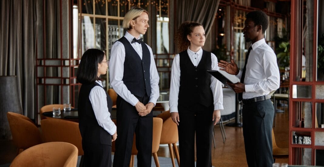 A group of four restaurant staff in uniforms—two women and two men—standing and discussing with a manager or supervisor inside a stylish dining area with tables and chairs.