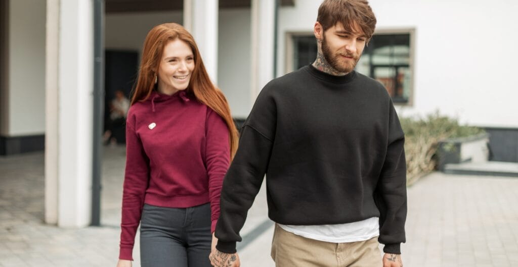 A couple walks hand in hand outdoors; the woman has long red hair and wears a burgundy hoodie, while the man has a beard and wears a black sweatshirt over a white tee.