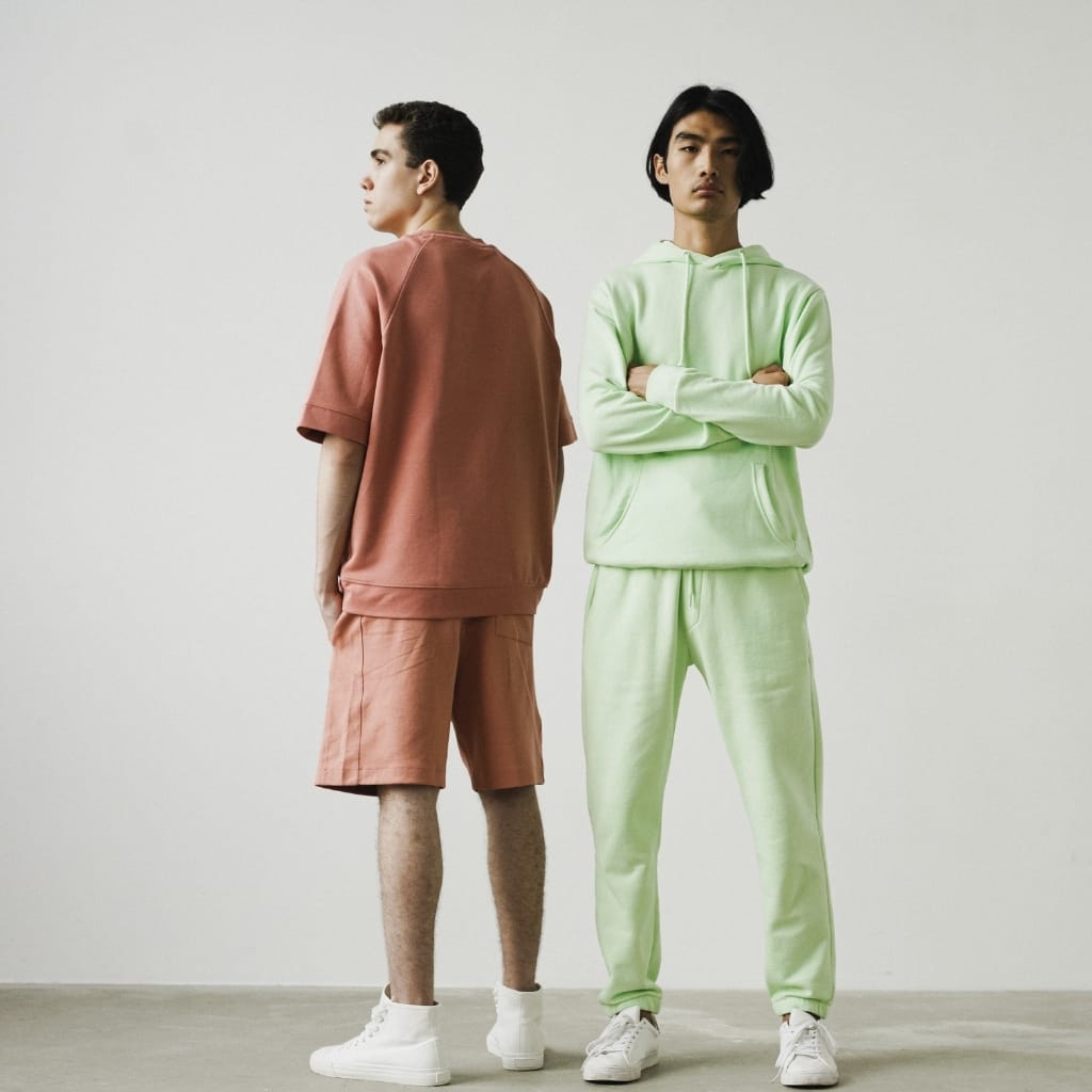Two men standing side by side against a light background. The man on the left wears a dusty rose oversized short-sleeve top and matching shorts, while the man on the right wears a mint green hoodie and sweatpants with white sneakers. Both have short black hair; one faces left with a neutral expression, the other faces forward with arms crossed.