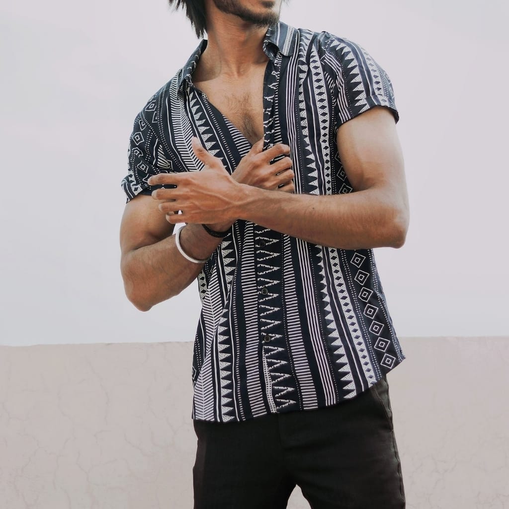 A man wearing a short-sleeve patterned shirt with geometric black-and-white stripes, standing against a light background, with one arm crossing over the other.