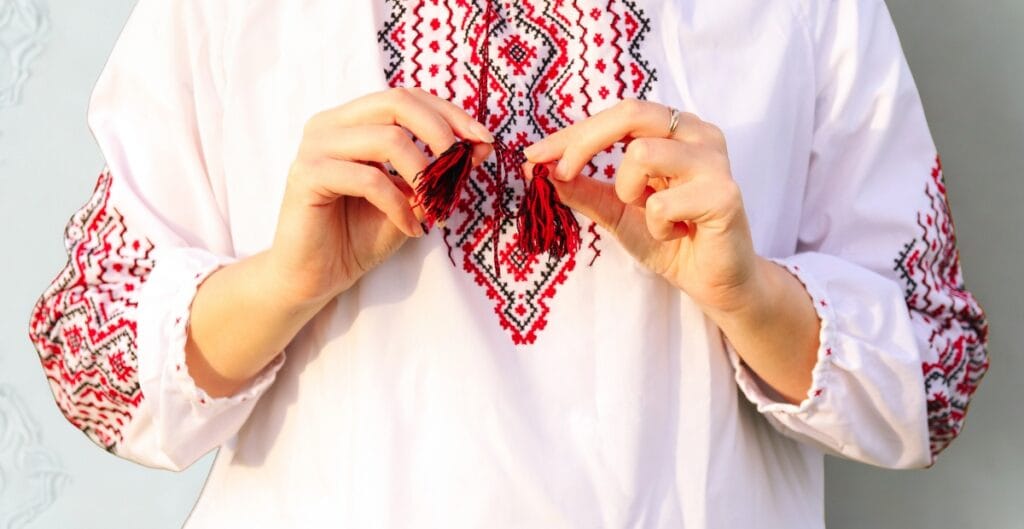 A person wearing a white embroidered blouse, holding a small red and black embroidered tassel with both hands.