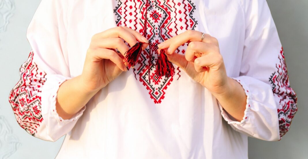 A person wearing a white embroidered blouse, holding a small red and black embroidered tassel with both hands.