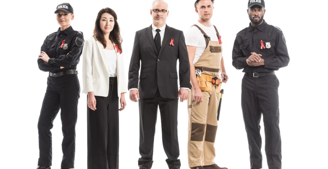A diverse group of five professionals standing side by side against a white background, including a police officer, a businesswoman, a businessman, a construction worker, and another police officer, all wearing red ribbons.