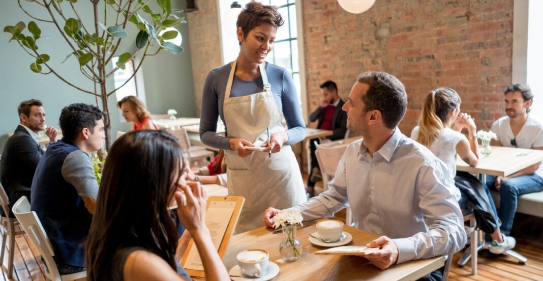 A bright cafe interior with several groups of people seated at wooden tables. In the foreground, a smiling server wearing a gray top and white apron chats with a man and a woman at a table with coffee cups and menus. Plants and a brick wall backdrop add warmth to the scene