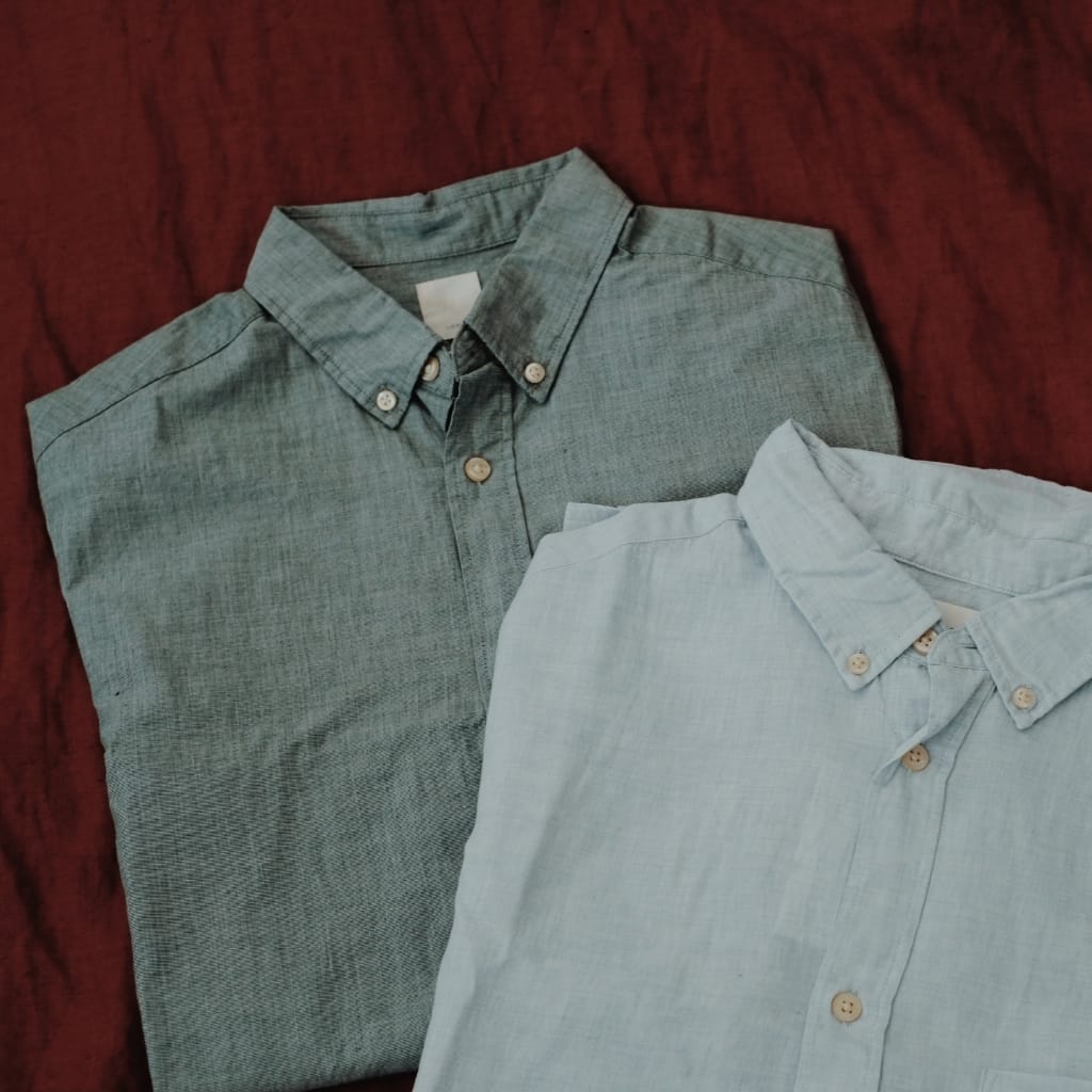Two button-down shirts in muted green and light blue laid flat on a maroon fabric background.