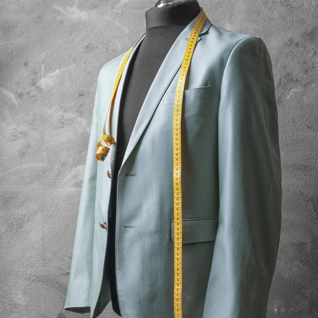 A light blue blazer on a mannequin, with a yellow measuring tape draped over the shoulder and torso, extending down the front. A rolled-up tape measure hangs from the lapel, and a gray textured background surrounds the display.
