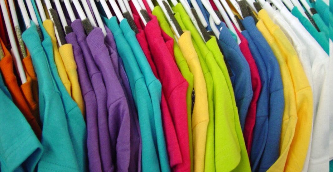 A close-up row of colorful folded T-shirts hanging on hangers, arranged in a rainbow spectrum from teal, turquoise, purple, pink, red, orange, yellow, green, to white.