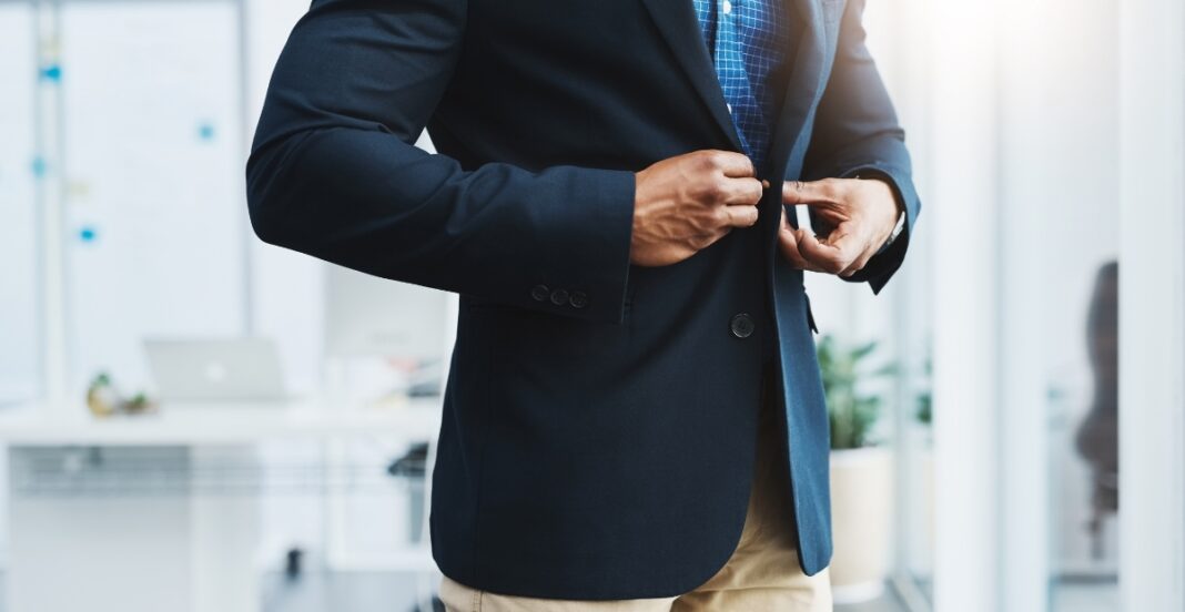 A person in a dark blazer buttoning up their shirt in an office setting.