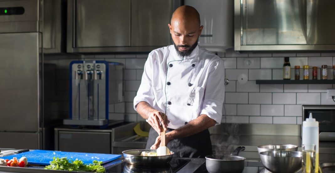 chef with a beard, dressed in a white chef's coat, is stirring food in a pan on a stovetop in a modern kitchen.