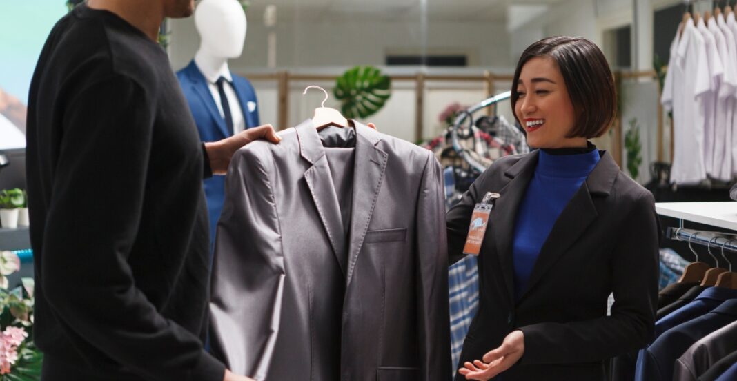 A woman with short hair wearing a black blazer and blue turtleneck smiling as she hands over a light grey blazer to a man at a clothing store.