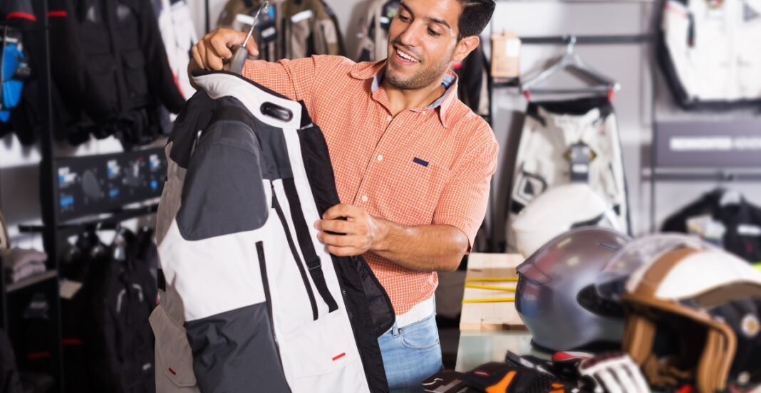A man is shopping for a backpack in a store that sells outdoor and sports gear. He is smiling and examining a large black and white backpack, with helmets and other gear on the counter beside him.