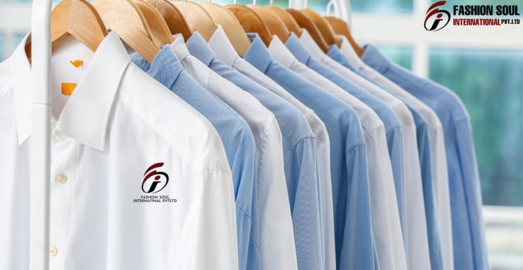 A row of white and light blue men's dress shirts hanging on wooden hangers on a rack. The shirts are displayed indoors with a bright, modern background. One white shirt in the foreground features a logo that reads 