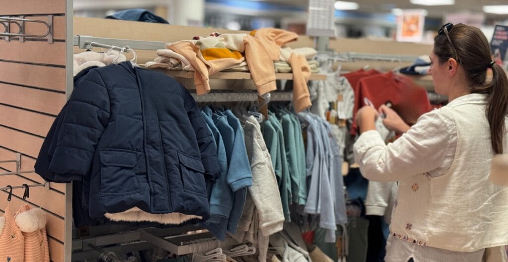 A woman shopping in a clothing store aisle, inspecting clothes on the rack. The aisle has children's jackets, sweaters, and other apparel hanging on the racks.