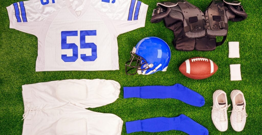 A collection of American football gear and accessories laid out on green turf, including a white jersey with the number 55, white pants, a blue helmet, blue socks, white shoes, a football, shoulder pads, two small white towels, and white wristbands.