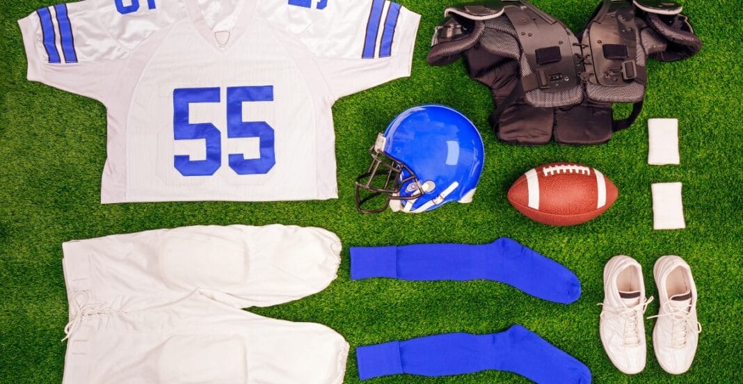 A collection of American football gear and accessories laid out on green turf, including a white jersey with the number 55, white pants, a blue helmet, blue socks, white shoes, a football, shoulder pads, two small white towels, and white wristbands.