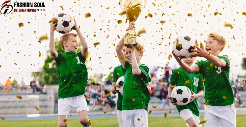Four young children in green soccer uniforms celebrating on a field, holding soccer balls and a trophy, with gold confetti falling around them. The background shows a blurred crowd and blue sky.