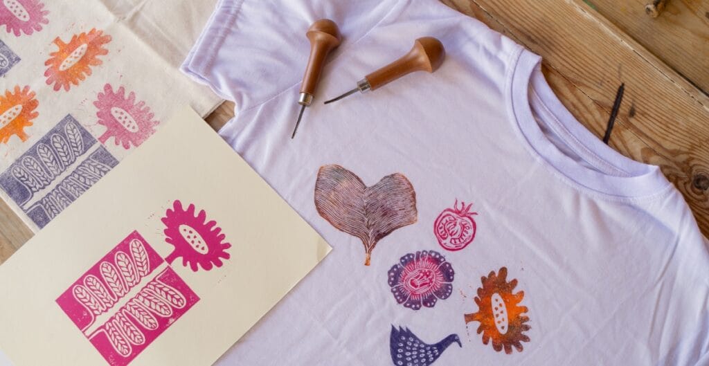 A white T-shirt spread on a wooden surface with several colorful, flower-shaped stamps printed on it. Nearby are stamping tools (handles and metal tips) and a cream-colored sheet with bold pink stamp designs. The scene suggests a DIY stamping or textile art project.